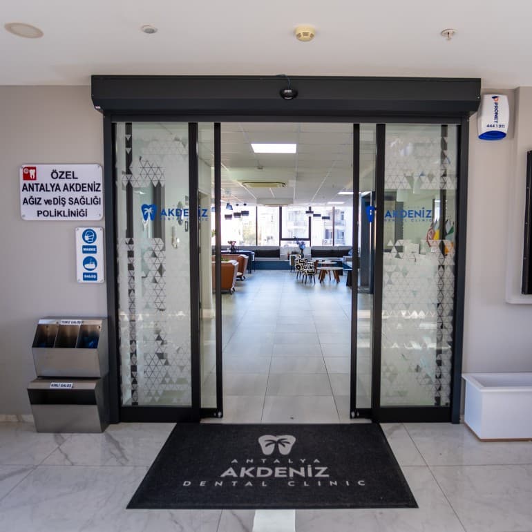 Wejście do kliniki Akdeniz Dental w Antalyi: szklane drzwi, tablica z nazwą placówki i wycieraczka z logo